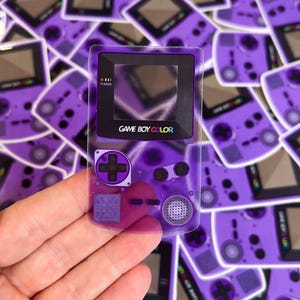 May include: A transparent purple sticker of a Game Boy Color handheld game console. The sticker has a black screen, buttons, and the text "GAME BOY COLOR". The background is filled with similar stickers.