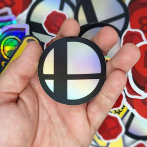 May include: A round, holographic sticker featuring the black and white Super Smash Bros. logo. The sticker has a glossy finish and is held in a hand, with other stickers visible in the background.