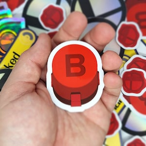 May include: A red sticker with a white border featuring a large, bold letter "B". The sticker has a 3D button design. Other stickers with a red seal design are in the background.