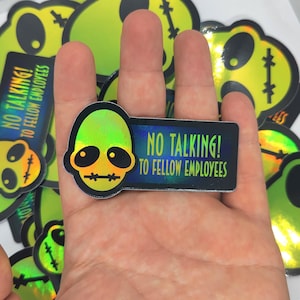Op de afbeelding: Een holografische sticker met een groene alienkop en de tekst "NO TALKING! TO FELLOW EMPLOYEES". De sticker heeft een zwarte rand en een rechthoekige vorm. Andere soortgelijke stickers zijn zichtbaar.