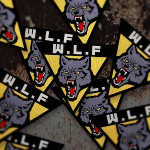 May include: A collection of patches featuring a snarling wolf head with red mouth and yellow eyes. The wolf is set against a yellow triangle, with the letters "W.L.F." in white on a black banner. The patches are scattered on a textured, rusty surface.