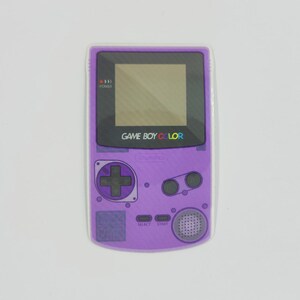 May include: A purple Game Boy Color handheld game console. The console has a black screen, a directional pad, and buttons labeled "SELECT" and "START". The text "GAME BOY COLOR" is printed on the console.