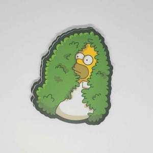 Puede incluir: Una pegatina troquelada que muestra a Homer Simpson asomándose por detrás de un arbusto verde. La cara amarilla y el pelo castaño de Homer son visibles, junto con su camisa blanca. La pegatina tiene un borde blanco y un contorno verde.
