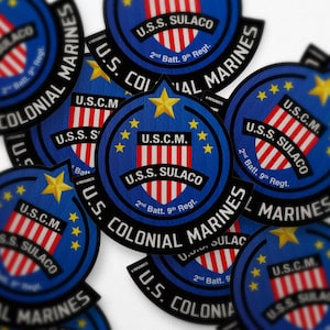 Pode incluir: Uma coleção de patches circulares com um design azul e vermelho com o texto "U.S.S. SULACO" e "U.S. COLONIAL MARINES". Cada patch inclui uma estrela e o texto "U.S.C.M.".