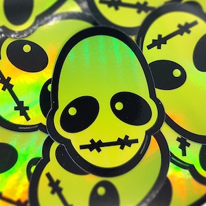 May include: A pile of holographic stickers featuring a lime green skull design with black outlines. The skull has large black oval eyes and a stitched mouth. The stickers have the text "UNUMSHOP" printed on them.