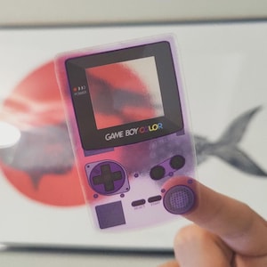 May include: A transparent purple Game Boy Color sticker with the text "GAME BOY COLOR". The sticker has a pink and purple gradient design and features the classic button layout, including a D-pad, A and B buttons, and start and select buttons.