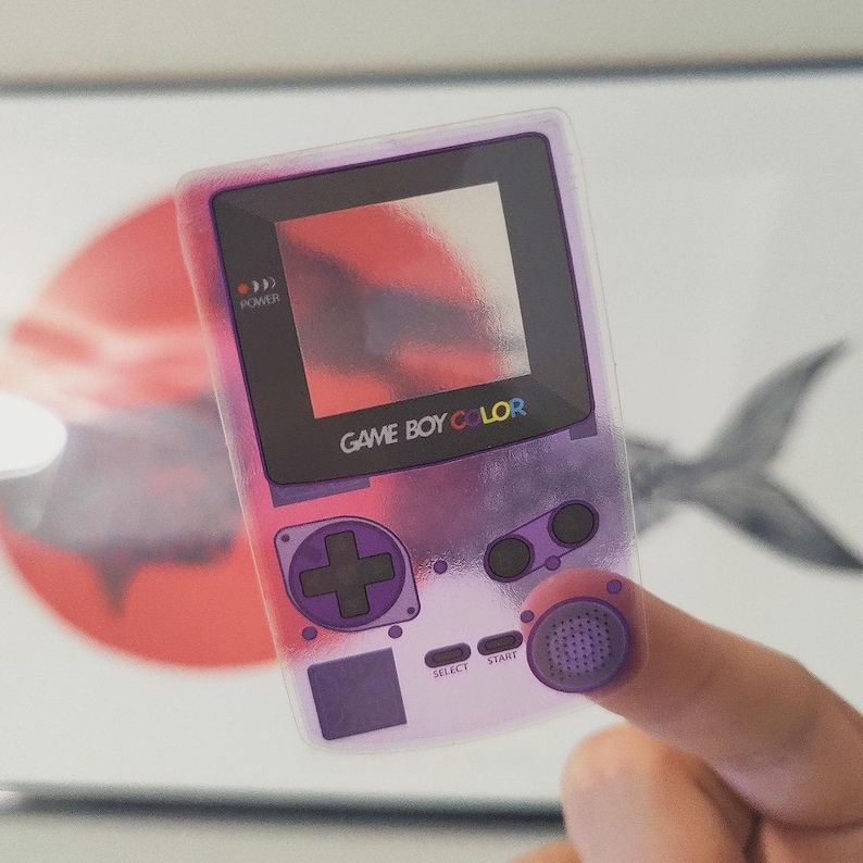 May include: A transparent purple Game Boy Color sticker with the text "GAME BOY COLOR". The sticker has a pink and purple gradient design and features the classic button layout, including a D-pad, A and B buttons, and start and select buttons.