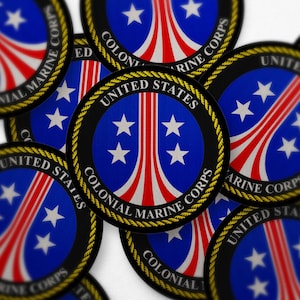 Pode incluir: Uma coleção de autocolantes redondos com um design azul e vermelho com estrelas brancas. O texto "UNITED STATES COLONIAL MARINE CORPS" está impresso à volta da borda. Os autocolantes têm uma borda preta e uma guarnição dourada em forma de corda.
