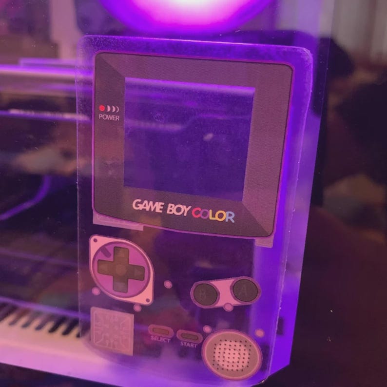May include: A transparent purple Game Boy Color handheld game console. The text "GAME BOY COLOR" is visible above the screen. The console features a directional pad, buttons, and the text "SELECT" and "START". The power indicator is lit.