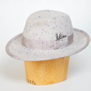 May include: A light gray fedora hat with blue paint splatters and a gray ribbon. The hat has a small feather accent and is sitting on a wooden hat form.