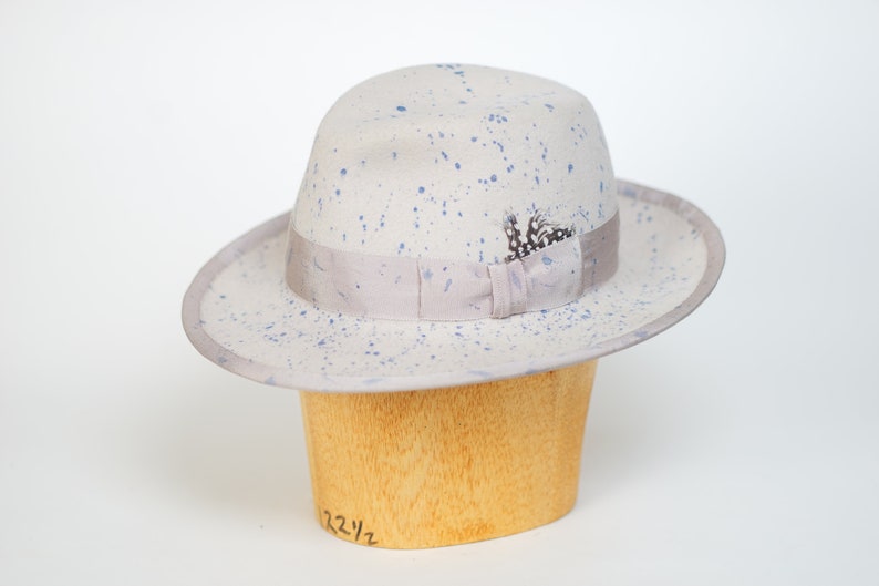 May include: A light gray fedora hat with blue paint splatters and a gray ribbon. The hat has a small feather accent and is sitting on a wooden hat form.