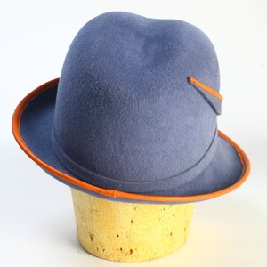 Puede incluir: Un sombrero fedora de fieltro azul con una banda y ribete de cuero marr&oacute;n. El sombrero tiene un ala ancha y una corona redondeada.