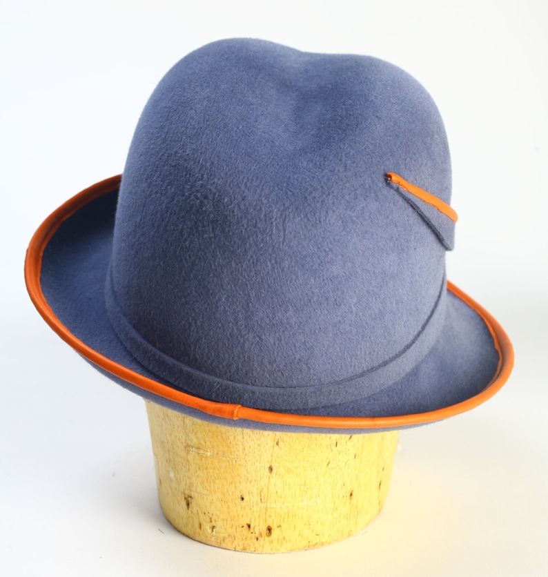 Puede incluir: Un sombrero fedora de fieltro azul con una banda y ribete de cuero marr&oacute;n. El sombrero tiene un ala ancha y una corona redondeada.