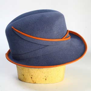 Puede incluir: Un sombrero de fieltro azul con un ala ancha y un ribete de cuero naranja. El sombrero est&aacute; sentado en una forma de sombrero de madera.