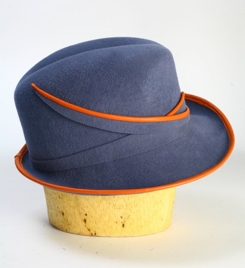 Puede incluir: Un sombrero de fieltro azul con un ala ancha y un ribete de cuero naranja. El sombrero est&aacute; sentado en una forma de sombrero de madera.
