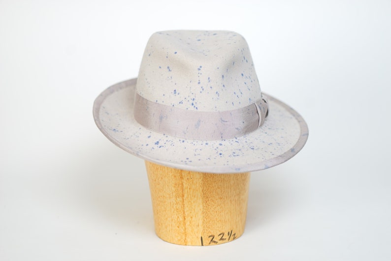 May include: A gray fedora hat with a light gray band and blue splatter paint. The hat is on a wooden hat form with the size "7 2 1/4" written on it.