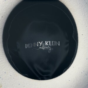 May include: The inside of a white felt hat with blue speckles. The hat has a black satin lining with the text "PENNY KLEIN millinery" embroidered in white.