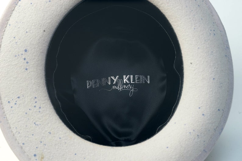 May include: The inside of a white felt hat with blue speckles. The hat has a black satin lining with the text "PENNY KLEIN millinery" embroidered in white.