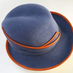 Puede incluir: Un sombrero fedora de fieltro azul con ribete de cuero naranja. El sombrero tiene una ala ancha y una corona ligeramente hundida.