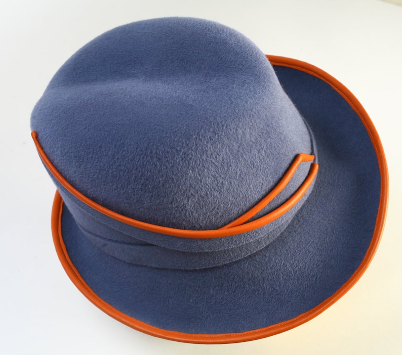 Puede incluir: Un sombrero fedora de fieltro azul con ribete de cuero naranja. El sombrero tiene una ala ancha y una corona ligeramente hundida.