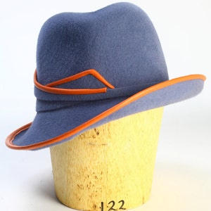 Puede incluir: Un sombrero de fieltro azul con un ala ancha y un ribete de cuero naranja. El sombrero est&aacute; sentado en una forma de sombrero de madera.