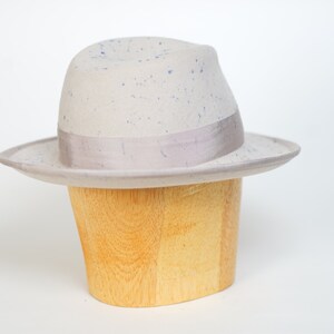 May include: A light gray fedora hat with a blue speckled pattern and a light gray ribbon band. The hat is displayed on a wooden hat stand.