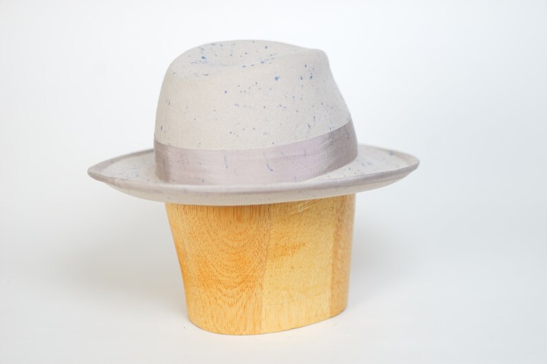 May include: A light gray fedora hat with a blue speckled pattern and a light gray ribbon band. The hat is displayed on a wooden hat stand.