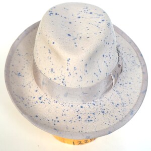 May include: A light gray fedora hat with a blue splatter paint design. The hat has a wide brim and a ribbon band around the crown.