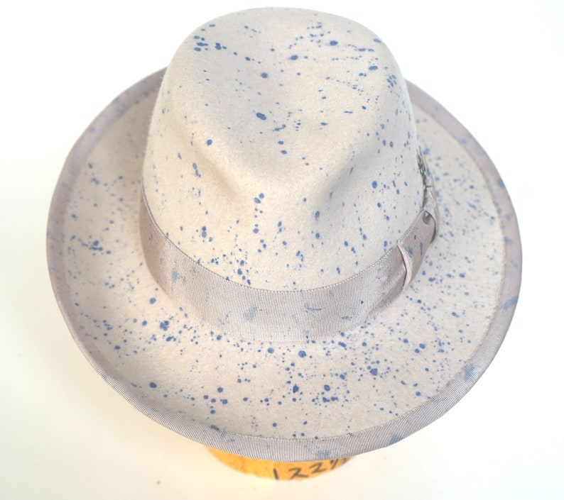 May include: A light gray fedora hat with a blue splatter paint design. The hat has a wide brim and a ribbon band around the crown.