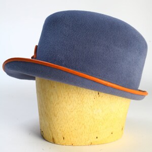 Puede incluir: Un sombrero de fieltro azul con una banda de cuero naranja alrededor del borde. El sombrero est&aacute; sentado en una forma de sombrero de madera.