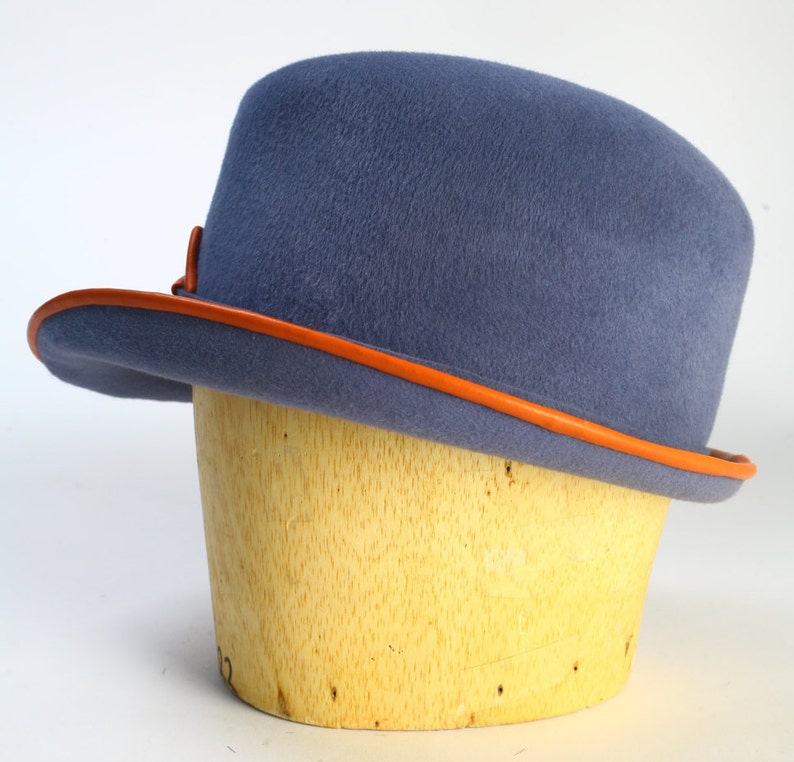 Puede incluir: Un sombrero de fieltro azul con una banda de cuero naranja alrededor del borde. El sombrero est&aacute; sentado en una forma de sombrero de madera.