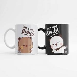 May include: Two ceramic mugs, one white and one black, with cartoon bear and panda illustrations. The white mug says "She's my Bubu" and the black mug says "He's my Dudu". Both mugs have a curved handle.