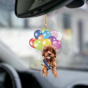 May include: A car charm featuring a brown puppy suspended by a bunch of colorful balloons. The puppy wears a blue ribbon and a medal. The balloons are in various colors, including red, blue, green, and yellow.