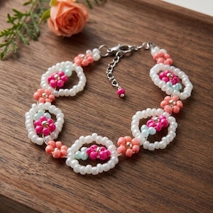 May include: A beaded bracelet featuring a series of circular white bead links, each containing a small flower motif made of bright pink and light blue beads. The bracelet includes a silver-toned metal chain extender with a small pink bead at the end.