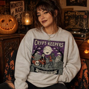 May include: A light gray crewneck sweatshirt featuring a graphic print of a band called The Crypt-Keepers. The illustration depicts a skeleton, a vampire, a werewolf, and a mummy performing music under a full moon with the text Monster Mash-Up.