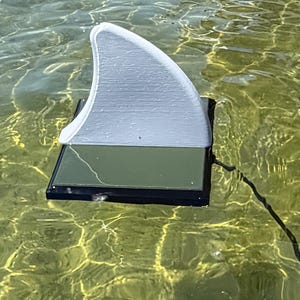 May include: A white shark fin-shaped object attached to a rectangular, dark-coloured base floating on the water surface. The base features a flat, dark green panel, and a thin black cord extends from the side of the unit into the surrounding water.