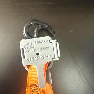 Könnte beinhalten: Nahaufnahme eines schwarzen Hakens und eines grauen Gurtschnappers, der an einem orangefarbenen Gurt befestigt ist. Der Gurtschnapper trägt die Aufschrift "RUEN DESIGN LLC", "V1-2M", "PATENT PENDING" und "STRAP SNAP".