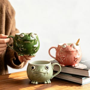 May include: Three ceramic frog-shaped mugs in green, pink, and light green. The mugs have large, expressive eyes and are designed with frog-like features. One mug is held by a person, while the others sit on a stack of books.
