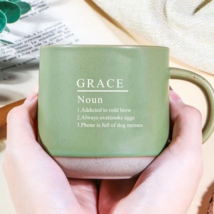 Könnte beinhalten: Eine salbeigrüne Keramiktasse mit hellbeigem Boden. Die Tasse trägt das Wort "GRACE" über dem Wort "Nomen" und drei humorvolle Aussagen. Die Tasse hat einen gebogenen Henkel und wird von zwei Händen gehalten.