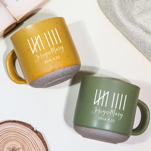 Pode incluir: Duas canecas de cerâmica, uma amarelo mostarda e outra verde oliva, com bases e alças cinzentas. Cada caneca tem um gráfico branco e o texto "Jerry❤Mary 2016.9.22". As canecas estão numa superfície branca.