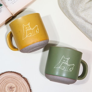 Puede incluir: Dos tazas de cerámica, una amarillo mostaza y otra verde oliva, cada una con una ilustración blanca de un gato leyendo un libro. Las tazas tienen una base gris moteada y asas a juego. Las tazas están sobre una superficie blanca.