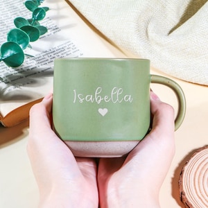 Pode incluir: Uma caneca de cerâmica verde com o nome "Isabella" em escrita branca e um pequeno coração branco. A caneca tem uma metade superior verde claro e uma metade inferior bege. A caneca é segurada por duas mãos.