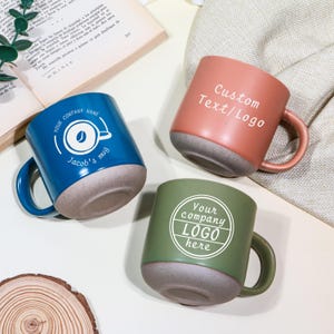 May include: Three ceramic mugs in blue, coral, and green. The blue mug has a coffee-themed logo and "Jacob's mug" text. The coral mug features "Custom Text/Logo". The green mug says "Your company LOGO here".