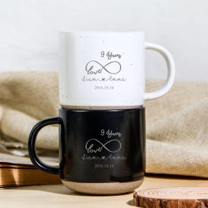 Pode incluir: Duas canecas de cerâmica empilhadas, uma branca e outra preta. A caneca branca tem um acabamento salpicado e a caneca preta tem um acabamento fosco. Ambas as canecas apresentam o texto "9 Anos", "Amor", nomes e uma data.