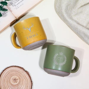 May include: Two ceramic mugs. One is mustard yellow with a deer antler design and the name "EMMA". The other is olive green with a laurel wreath and the letter "T". Both mugs have a speckled grey base and matching handles.