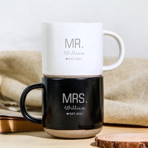 Puede incluir: Dos tazas de cerámica apiladas, una blanca y otra negra. La taza blanca tiene el texto "MR. William ♥ EST.2025". La taza negra tiene el texto "MRS. William ♥ EST.2025". Ambas tazas tienen un diseño moteado y un asa redondeada.