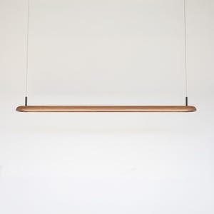 May include: A modern, minimalist pendant light fixture. The light features a long, rectangular wooden body with rounded ends, suspended by thin wires. The wood has a warm, natural tone. The fixture is designed to provide ambient lighting.
