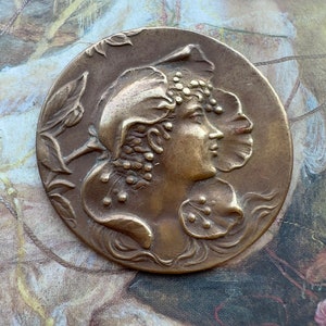 May include: A bronze-colored round medallion featuring a profile of a woman with floral and berry accents in her hair and on her head covering. The medallion is set against a backdrop of a vintage painting with muted colors.