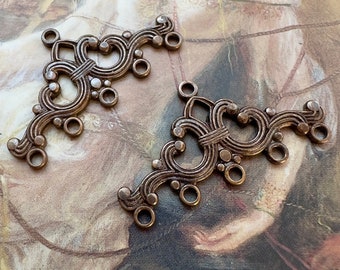 May include: Two antique gold-toned metal decorative pieces with intricate scrollwork and multiple loops. The pieces are similar in design and have a vintage aesthetic.