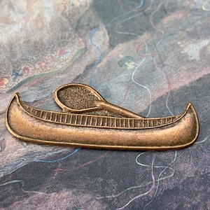 May include: A bronze-colored metal canoe pin with a paddle. The canoe has detailed lines and a curved shape. The paddle rests on top of the canoe. The background is a patterned fabric with muted colors.